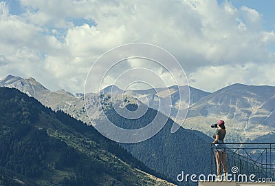 A female photographer in a red cap with a camera stands on the balcony opposite greek mountains and village in Greece Stock Photo