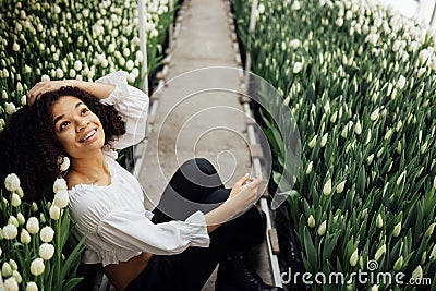 Female portrait of attractive darkskinned girl sitting on path among tulips in greenhouse Stock Photo