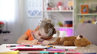 Female preschooler drawing picture, teddy lying on table, happy childhood, hobby Stock Photo
