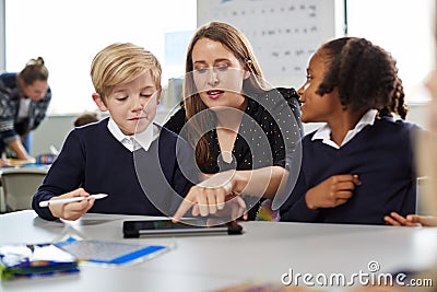 Female school teacher helping two kids using a tablet computer at desk in a primary school classroom, front view Stock Photo
