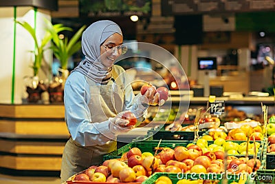 Female seller in hijab browsing and checking apples in supermarket, woman in apron smiling at work in store in fruit and Stock Photo