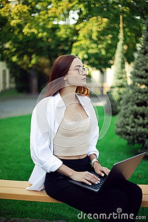 A female student in glasses sits on a bench with laptop. Freelance work in the fresh air Stock Photo