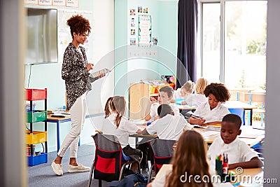 Female Teacher With Digital Tablet Teaches Group Of Uniformed Elementary Pupils In School Classroom Stock Photo