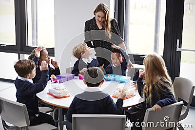 Female teacher stands talking to a group of primary school kids sitting together at a round table eating their packed lunches Stock Photo