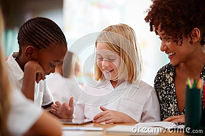 Female Teacher With Two Elementary School Pupils Wearing Uniform Using Digital Tablet At Desk Stock Photo