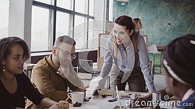 Female team leader standing near table and giving direction to young creative team. Brainstorming of multiethnic group. Stock Photo