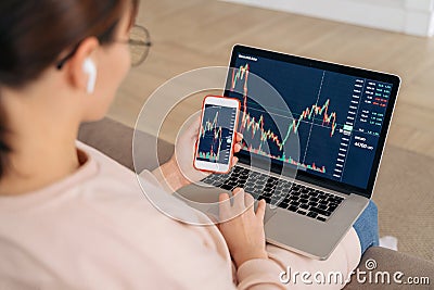 Female trader investor analyst using financial stock market app on modile phone and laptop at home Stock Photo