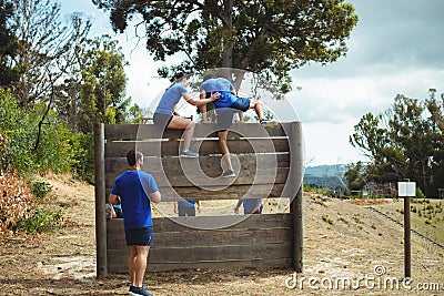 Female trainer assisting fit man to climb over wooden wall during obstacle course Stock Photo