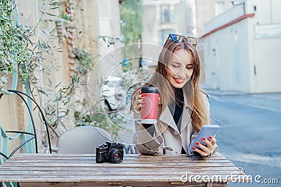 Female traveler chatting, sharing files, resting in cafe terrace table with vintage camera and coffee in reusable cup Stock Photo