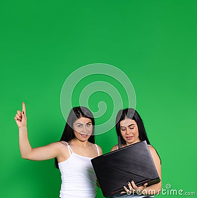 Female twins on a green background. Holding a laptop, points finger up to the empty space. Technology and advertisment Stock Photo