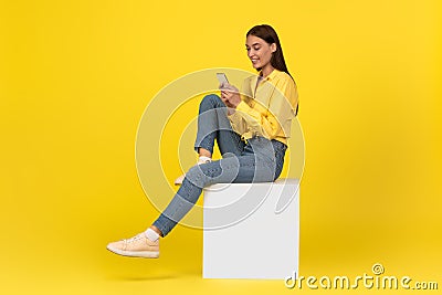 Female Using Smartphone Texting Sitting On Cube Over Yellow Background Stock Photo