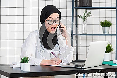 Female veiled scientist sitting on her desk looking at laptop opens her mouth with shocked face emotion while talking on Stock Photo