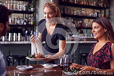 Female Waitress Serving Food To Romantic Couple Sitting At Restaurant Table Stock Photo