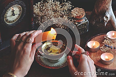 Female wiccan witch wearing vintage jewelry holding yellow candle and pouring wax into a red white gold colored vintage teacup Stock Photo