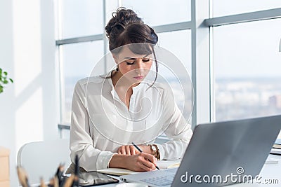 Female writer working in light modern office, writing down new ideas her notebook, searching information using portable Stock Photo