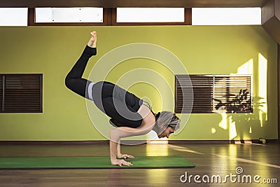 A female yoga practitioner doing a variation of the Eka Pada Bakasana exercise, handstand with leg extended, exercising on a mat Stock Photo