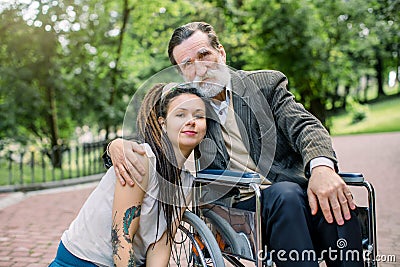 Female young social worker and disabled man on a walk. Girl and grandfather sitting in a wheelchair, hugging and posing Stock Photo