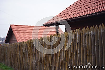 Fence made of sharp wooden stakes, wooden houses with red tiled roof and cloudy sky Stock Photo