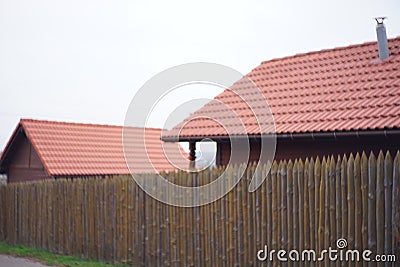 Fence made of sharp wooden stakes. Wooden houses with red tiled roof and sky. Unfocused image Stock Photo