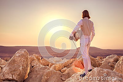 Fencer man standing on top of the rock holding fencing mask and a sword on sunset background Stock Photo