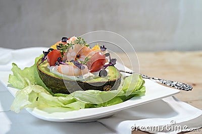 Filled avocado with shrimps, tomatoes, mayonnaise and cress garnish on lettuce and a white plate, light rustic background, festive Stock Photo