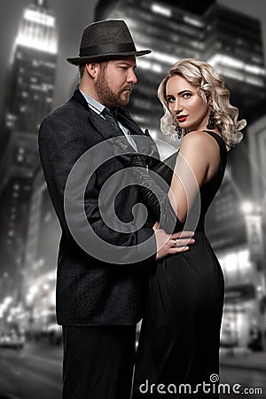 Film noir. Detective man in a raincoat and hat and a dangerous woman with red lips in black dress. Couple stands against Stock Photo