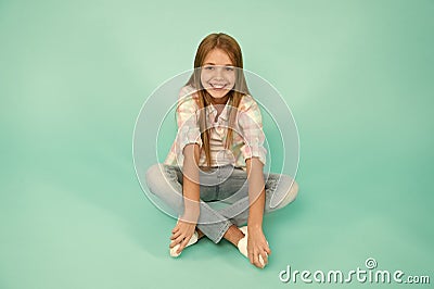Find place to relax. Girl happy face sit on floor attentive looking at camera turquoise background. Kid girl with long Stock Photo