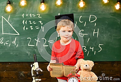 First former interested in studying, education. Kid boy in graduate cap holds book in classroom, chalkboard on Stock Photo