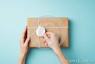 First person top view photo of girl`s hands holding pricetag on craft paper giftbox with small white bow on isolated pastel blue Stock Photo