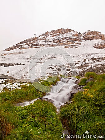 First snow in Alps touristic region. Fresh green meadow with rapids stream. Peaks of Alps mountains in background. Stock Photo