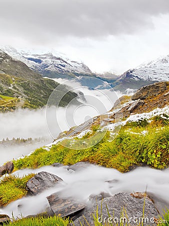 First snow in Alps touristic region. Fresh green meadow with rapids stream. Peaks of Alps mountains in background. Stock Photo