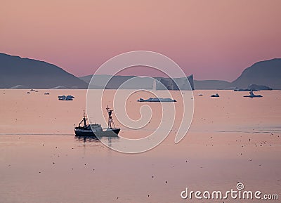 Fish trawler among huge icebergs during midnight sun with sunset and sunrise on the horizon. Fisher boat in arctic ocean Stock Photo