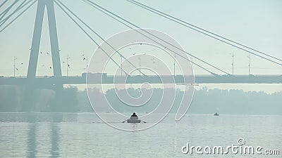 Fisherman sailing under big bridge on inflatable boat, misty weather, big city Stock Photo