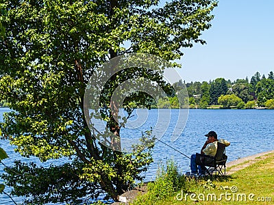 Fisherman sits alone in shade of tree beside water patiently waiting for fish to bite. Editorial Stock Photo