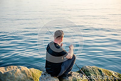 A fisherman sits on a rock by the sea with a fishing rod at sunset. Single sea fishing. Day at the beach. Copy space for text Stock Photo