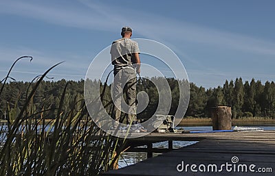 Fisherman on wooden pier catching fishes on reel rod, standing in summer near lake Editorial Stock Photo