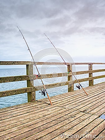 Fishers rods against handrail of wooden bridge. Fishing on harbor mole. Overcast day Stock Photo