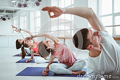 Fit adult women and man practicing yoga poses in fitness class. Group of healthy strong people doing fit exercises in white gym Stock Photo