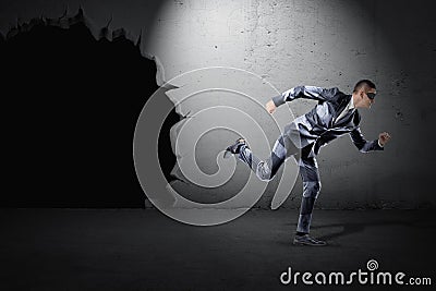 A fit young businessman in a striped suit and a black mask running away from a black hole in a wall. Stock Photo