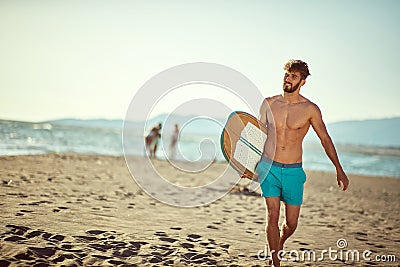 Fit young beardy male walking on the sunny beach, holding surfboard, smiling. Copy space Stock Photo