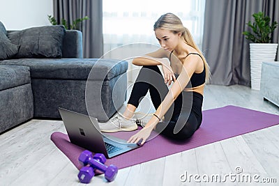 Fit young woman watching at online workout videos on laptop while sitting on exercise mat in living room at home Stock Photo