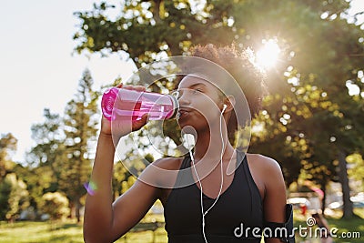 Fitness athlete young african american woman listening to music on earphones drinking water in a reusable water bottle Stock Photo