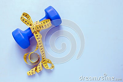 Fitness and weight loss concept. Blue dumbbell with bow of yellow centimeter on a blue background. Top view. Stock Photo