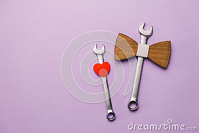 Flat lay composition with wooden bow tie, little red heart, two combination wrenches on pastel violet background with copy space. Stock Photo