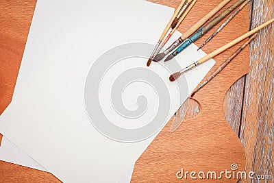 Flat lay of white Papers, paint brushes and wooden palette on brown wooden table. Stock Photo