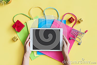 Flat lay of woman hands using tablet with miniature gift boxes. trolley and colorful bags on yellow background. Top view and copy Stock Photo