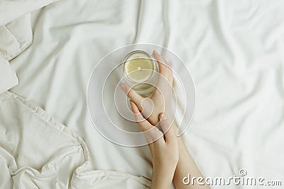 Flatlay of woman`s hands holding glass in lemon water in bed on white sheets Stock Photo