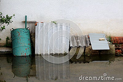 Flooded backyard junk covered with partially rusted metal barrel and grey roof tiles leaned on family house wall Stock Photo