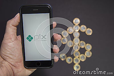Florianopolis, Brazil. 07/10/2020: Man hand holding smartphone with PIX logo with dollar sign made by 1 Real coins on background. Editorial Stock Photo