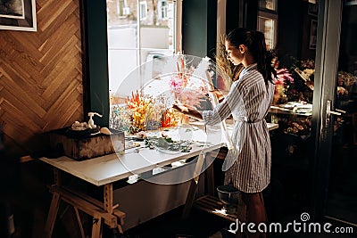 Florist dressed in a striped dress stands near the table with colorful dried flowers and holds transparent packaging Stock Photo